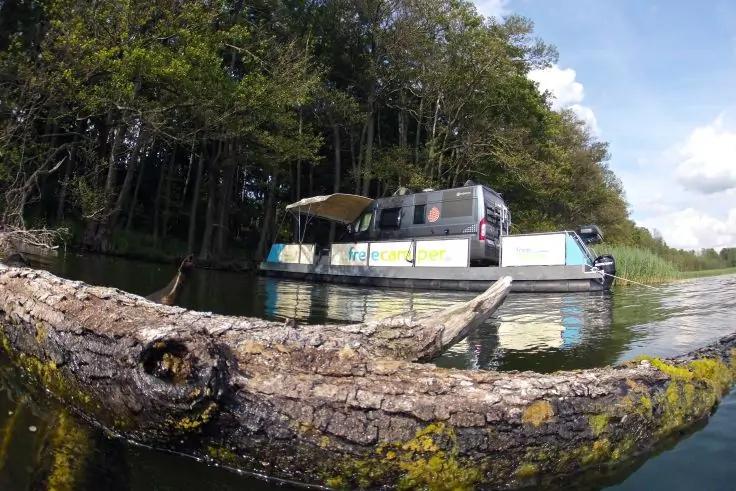 Im Vordergrund ein teilweise im Wasser liegender, moosbedeckter Baumstamm; dahinter eine flache Bootsplattform mit einem geparkten Van, am Ufer Wald und bewölkter Himmel