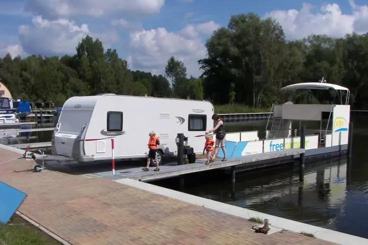 Weißer Wohnwagen auf einer Fahrzeugfähre am Anleger; zwei Personen mit orangefarbenen Schwimmwesten stehen daneben; Ufer, Bäume und teilweise bewölkter Himmel im Hintergrund