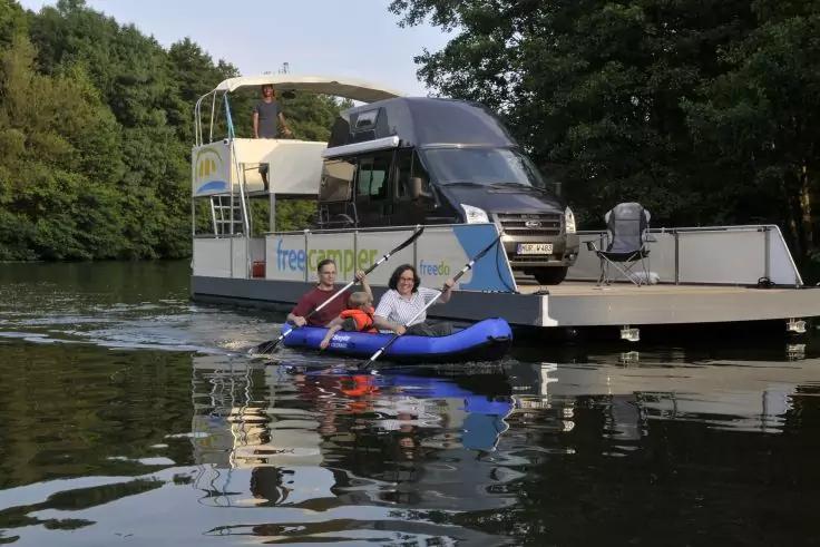 Zwei Personen in blauem Schlauchkajak paddeln vor einem kleinen Ausflugsboot mit einem schwarzen Wohnmobil auf dem Deck, baumbestandene Uferlinie im Hintergrund