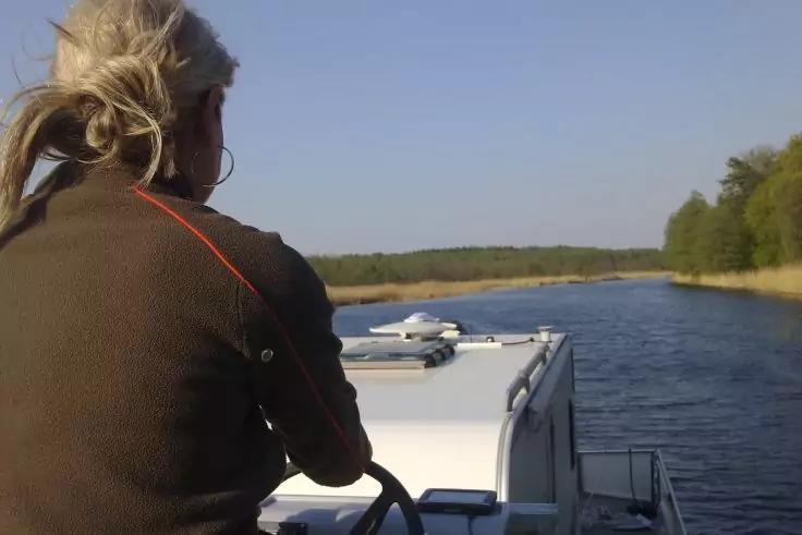 Person mit blondem Zopf von hinten am Steuer eines Boots, Fluss mit Schilf und Bäumen, blauer Himmel