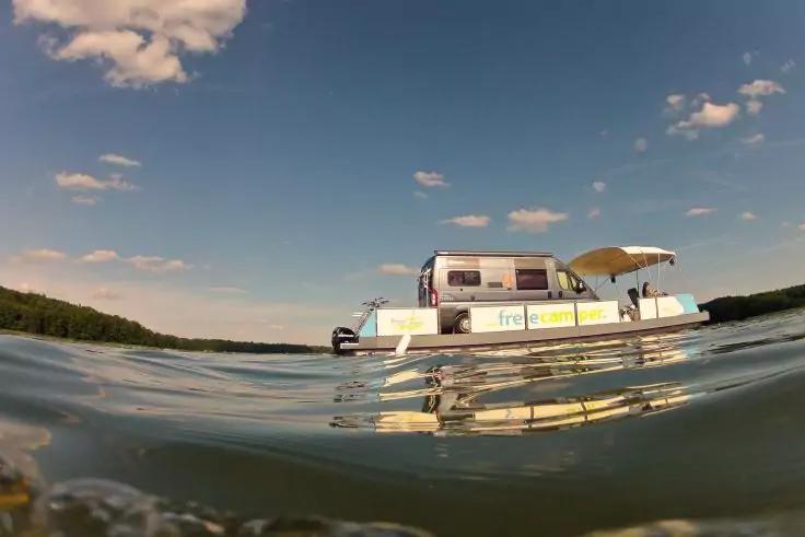 Pontonboot mit Verdeck und Beschriftung auf ruhigem See, niedrige Perspektive teilweise im Wasser, Spiegelung im Wasser, bewaldetes Ufer im Hintergrund, blauer Himmel mit vereinzelten Wolken