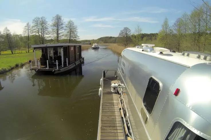 Silbernes Boot am Holzsteg, links ein dunkles Hausboot, in der Ferne ein kleines Boot, Ufer mit Bäumen, blauer Himmel mit wenigen Wolken