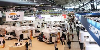 Halle mit mehreren ausgestellten Wohnmobilen, laufenden Besuchern und h&auml;ngenden Schildern mit Schriftzug 'carado'