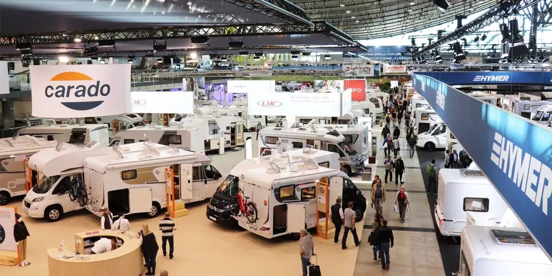 Halle mit mehreren ausgestellten Wohnmobilen, laufenden Besuchern und h&auml;ngenden Schildern mit Schriftzug 'carado'