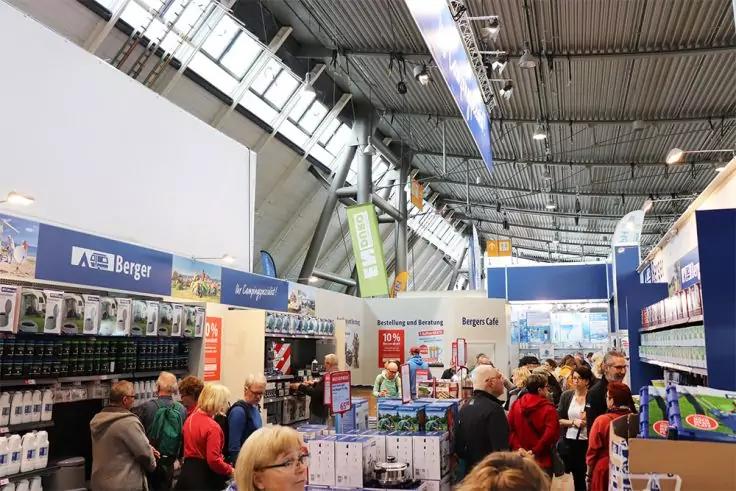 Innenraum mit hoher Decke, h&auml;ngenden Bannern, Reihen von St&auml;nden und Regalen, Tischen mit Produkten und Personen