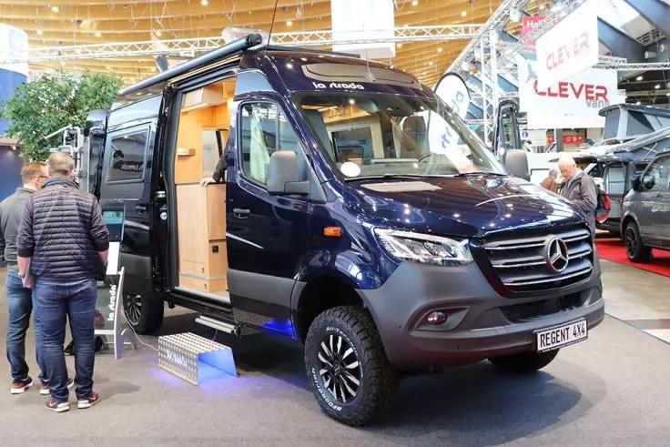 Dunkelblauer Mercedes-Camper in einer Messehalle, ge&ouml;ffnete Seitent&uuml;r mit sichtbarer Holzinneneinrichtung, mehrere Personen stehen daneben