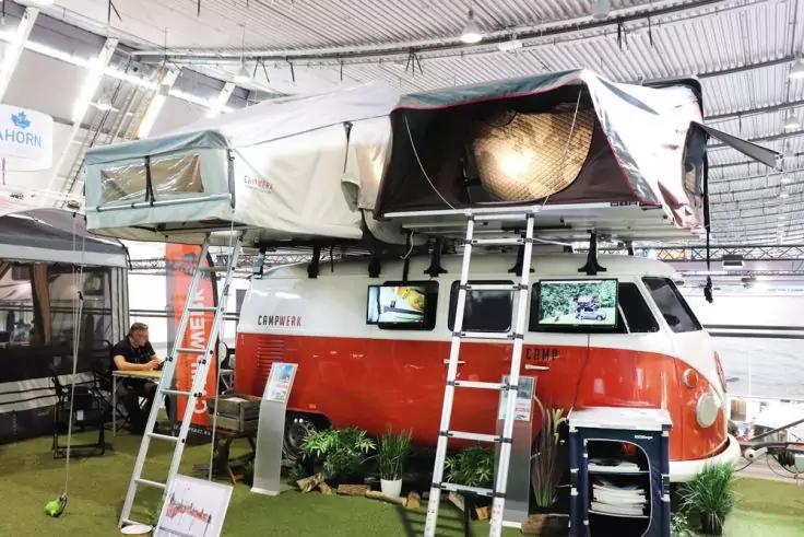 Roter-wei&szlig;er Campingbus mit aufgeklapptem Dachzelt, zwei Leitern, Bildschirme an der Seite, Infostand, Pflanzen und eine sitzende Person in einer Messehalle