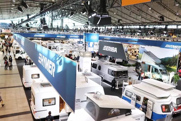 Gro&szlig;e Ausstellungshalle mit mehreren wei&szlig;en Wohnmobilen, blauen h&auml;ngenden Bannern mit Schriftzug und Personen im Hintergrund