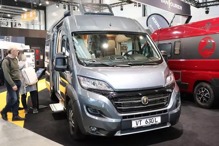 Grauer Camper-Van von vorne mit ge&ouml;ffneter T&uuml;r und ausgefahrener Trittstufe, in einer Halle neben einem roten Van, zwei Personen links