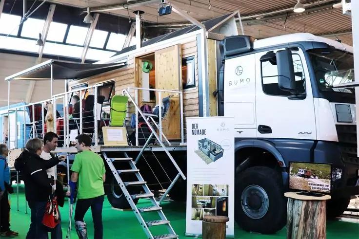 Wei&szlig;er Lastwagen in einer Halle mit seitlich ge&ouml;ffnetem Ausstellungsaufbau, Metalltreppe, Holzelementen und mehreren Personen im Vordergrund