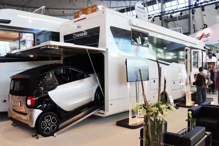 Wei&szlig;es Wohnmobil mit ge&ouml;ffnetem Heck, in dem ein kleiner zweifarbiger Kleinwagen steht; Personen stehen daneben, im Hintergrund weitere Fahrzeuge und Hallendecke