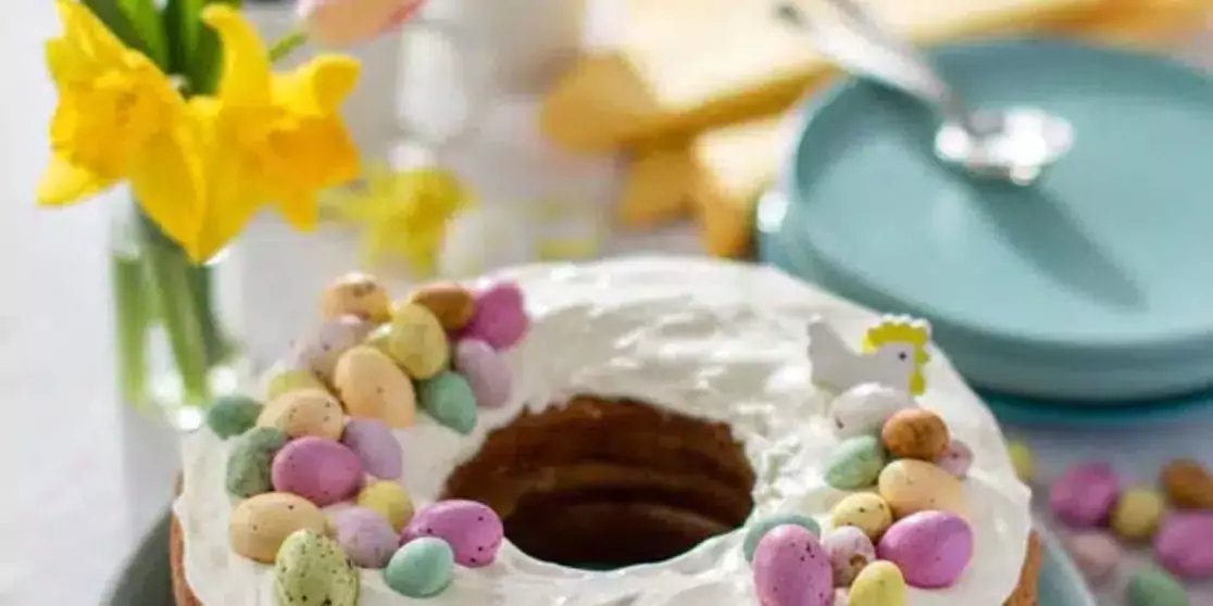 Runder Kuchen mit wei&szlig;er Glasur, dekoriert mit kleinen, gesprenkelten bunten Schokoladeneiern; gelbe Narzissen im Hintergrund