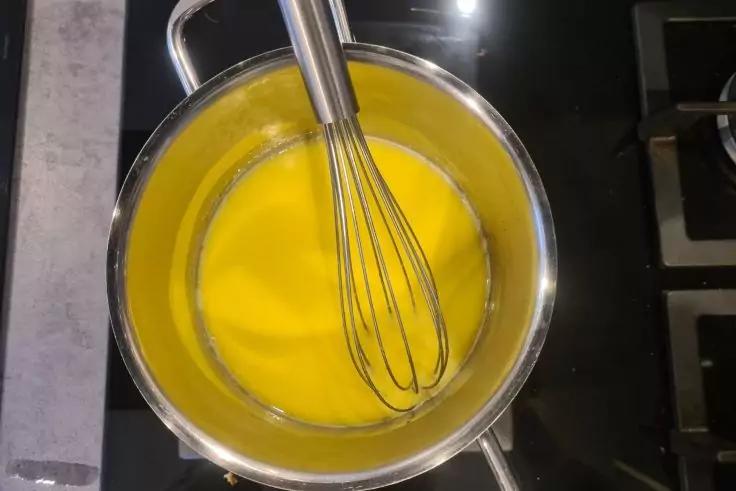 Edelstahltopf mit gelber Fl&uuml;ssigkeit und Schneebesen auf schwarzem Kochfeld