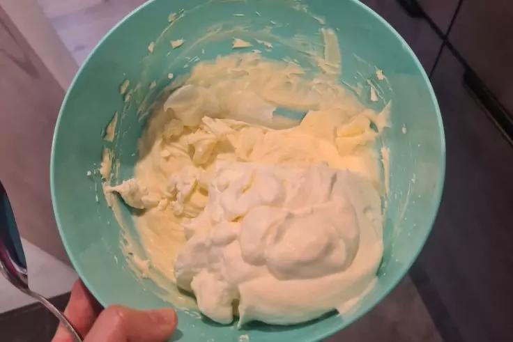 T&uuml;rkise R&uuml;hrsch&uuml;ssel mit wei&szlig;er cremiger Masse, Hand h&auml;lt die Sch&uuml;ssel, K&uuml;chenboden und Schrank im Hintergrund