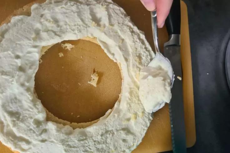 Hand mit L&ouml;ffel &uuml;ber wei&szlig;er Creme am Rand eines runden Tortenbodens mit Loch in der Mitte auf einem braunen Brett