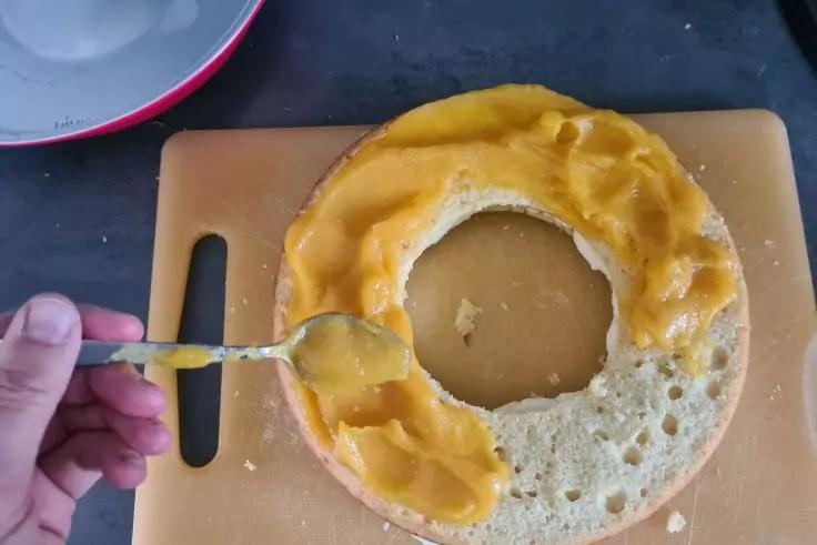 Hand mit L&ouml;ffel streicht gelbe Creme auf einen ringf&ouml;rmigen Biskuitkuchen auf einem Schneidebrett; Sch&uuml;ssel mit Creme links oben sichtbar