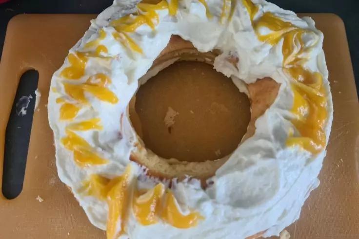 Runder Kuchen mit Loch in der Mitte, bedeckt mit wei&szlig;er Creme und mit gelber So&szlig;e betr&auml;ufelt, auf einem Schneidebrett