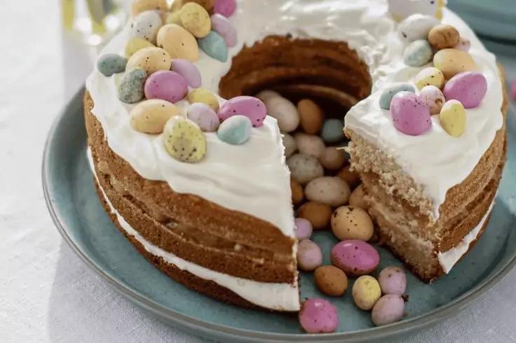 Runder Kuchen mit wei&szlig;er Glasur, ein St&uuml;ck herausgeschnitten, hohler Kern gef&uuml;llt mit pastellfarbenen Schokoladeneiern; einige Eier auf der Glasur und auf einem blauen Teller