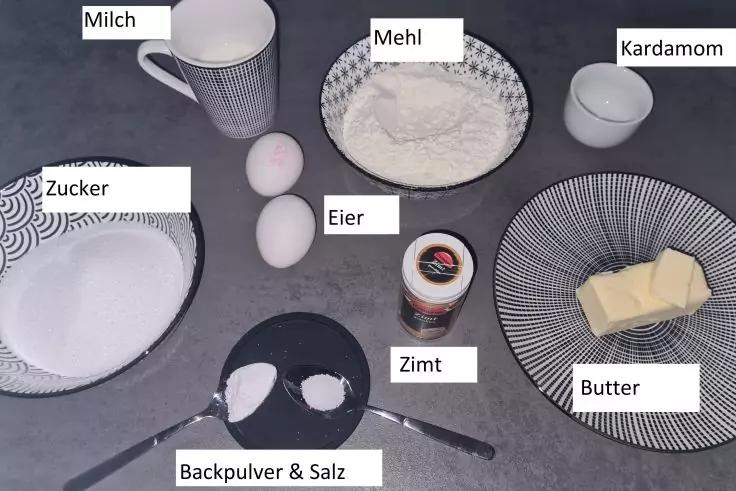 Zutaten auf dunkler Arbeitsfl&auml;che: Tasse mit Milch, Schale mit Mehl, kleine Schale mit Kardamom, Teller mit Zucker, zwei Eier, kleines Glas Zimt, zwei Messl&ouml;ffel mit Backpulver und Salz, Teller mit einem St&uuml;ck Butter
