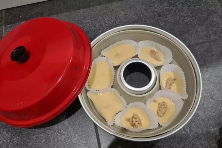 Runde Metall-Backform mit Rohr in der Mitte, sieben wei&szlig;e Papierf&ouml;rmchen mit hellgelben St&uuml;cken, einige mit braunen Flecken; roter Deckel daneben auf grauer Arbeitsfl&auml;che
