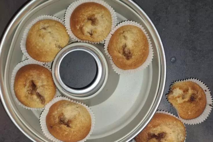 Sieben Muffins in wei&szlig;en Papierf&ouml;rmchen, f&uuml;nf in einer runden Backform mit Loch in der Mitte, zwei neben der Form auf dunkler Oberfl&auml;che
