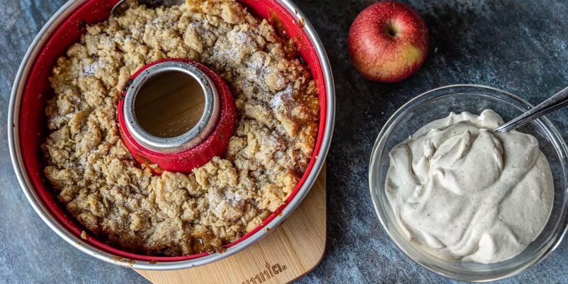 Runde rote Kuchenform mit kr&uuml;meliger Oberfl&auml;che auf einer Holzunterlage, daneben eine ganze rote Apfel und eine Glassch&uuml;ssel mit wei&szlig;er Creme und einem L&ouml;ffel auf dunklem Untergrund