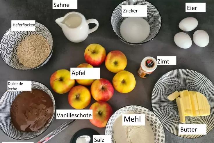 Mehrere &Auml;pfel, Schalen mit Haferflocken, Zucker, Mehl und Dulce de Leche, kleine Kanne mit Sahne, Butterst&uuml;cke, drei Eier, Glas mit Zimt, kleine Schale mit Salz und eine Vanilleschote auf dunklem Untergrund