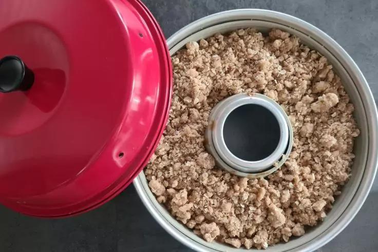 Runde Metallform mit zentraler R&ouml;hre, gef&uuml;llt mit kr&uuml;meliger Masse; roter Deckel daneben auf grauem Untergrund