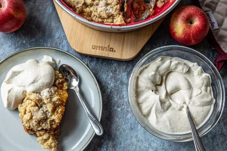 Aufsicht auf eine runde Backform mit Streuseln und einer geschnittenen Fruchth&auml;lfte, ein Teller mit einer streuselbedeckten Portion und einem L&ouml;ffel, eine Sch&uuml;ssel mit hellem cremigem Aufstrich und zwei ganze rote &Auml;pfel auf grauem Untergrund