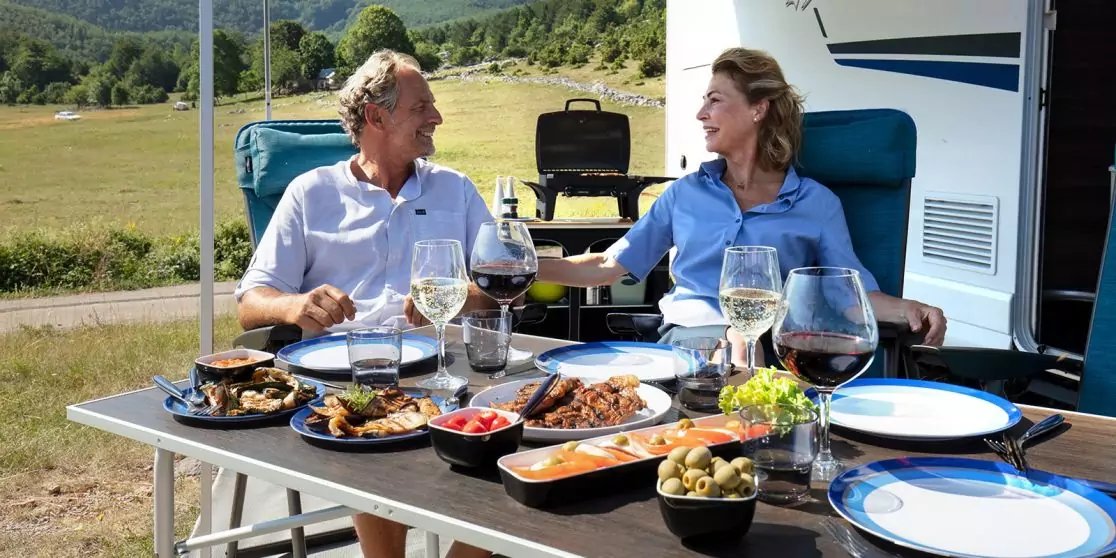 Zwei Personen sitzen an einem Campingtisch vor einem Wohnmobil. Auf dem Tisch stehen Teller und Schalen mit Speisen sowie mehrere Gl&auml;ser; im Hintergrund Wiese und Berge