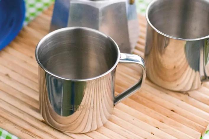 Zwei Edelstahlbecher auf einer Holzlattenunterlage, einer vorne mit Henkel, im Hintergrund ein Teil einer Metallkanne