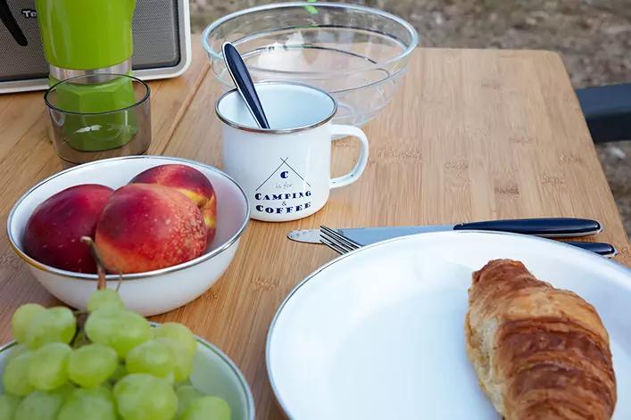 Holztisch mit Croissant auf wei&szlig;em Teller, wei&szlig;e Tasse mit Aufdruck 'Camper Coffee' und L&ouml;ffel, Sch&uuml;sseln mit Pfirsichen und gr&uuml;nen Trauben, Glassch&uuml;ssel, gr&uuml;ner zylindrischer Beh&auml;lter, Messer und Gabel