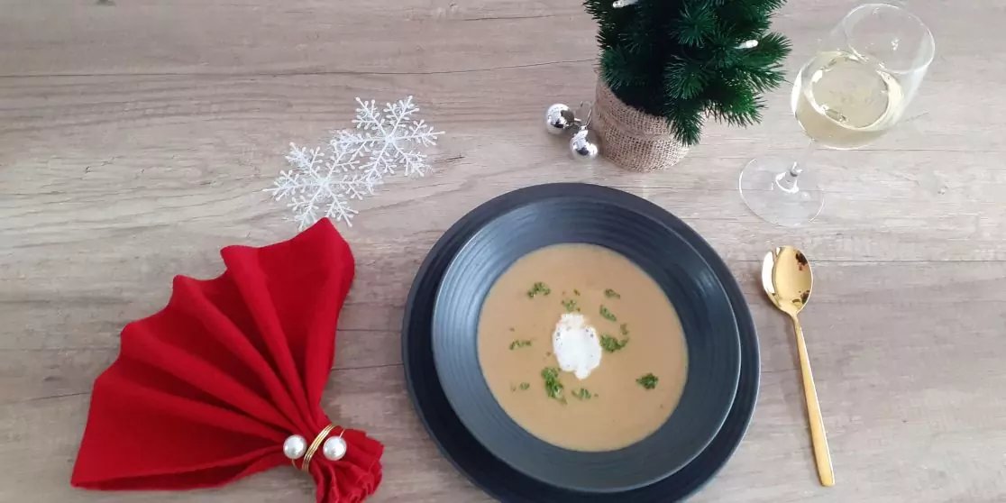 Schwarze Sch&uuml;ssel mit heller Suppe, in der Mitte Klecks wei&szlig;e Creme und gehackte Kr&auml;uter; goldener L&ouml;ffel rechts; rote, gefaltete Stoffserviette mit silbernen Gl&ouml;ckchen links; wei&szlig;e Schneeflocken-Dekoration oben links; kleiner gr&uuml;ner Baum mit silbern