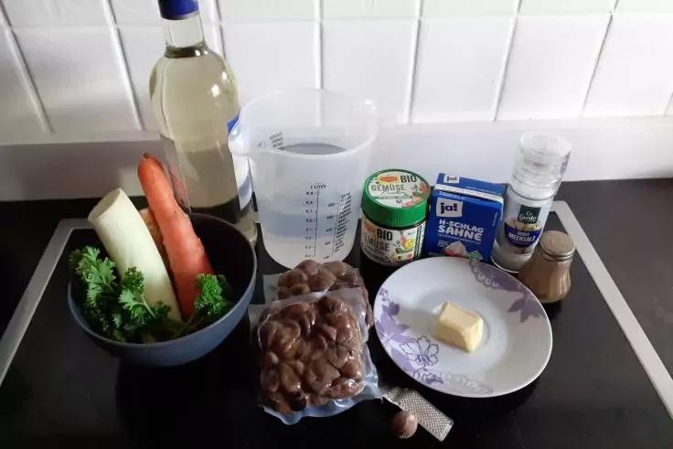 K&uuml;chenarbeitsfl&auml;che vor wei&szlig;er gefliester Wand mit Messbecher mit Wasser, Flasche mit heller Fl&uuml;ssigkeit, Sch&uuml;ssel mit Lauch, Karotte und Petersilie, Plastikt&uuml;te mit braunen Bohnen, kleiner Teller mit St&uuml;ck Butter, Knoblauchzehe, Milchkarton, Creme-/