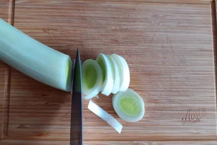 Holzschneidebrett mit einer Lauchstange; ein Messer schneidet runde Lauchscheiben