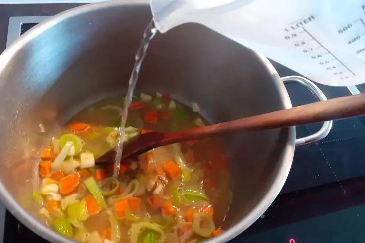 Topf auf dem Herd mit gew&uuml;rfeltem Gem&uuml;se (Karotten, Lauch, Zwiebeln), Holzl&ouml;ffel und Wasser, das aus einem Messbecher eingegossen wird