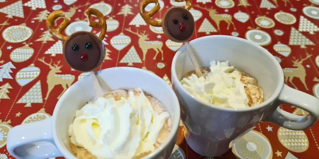 Zwei wei&szlig;e Tassen mit Getr&auml;nken, jeweils mit Schlagsahne und Kakaopulver, auf rotem Tischtuch mit Weihnachtsmotiven; in jeder Tasse ein Rentier-Stecker mit Knopfgesicht