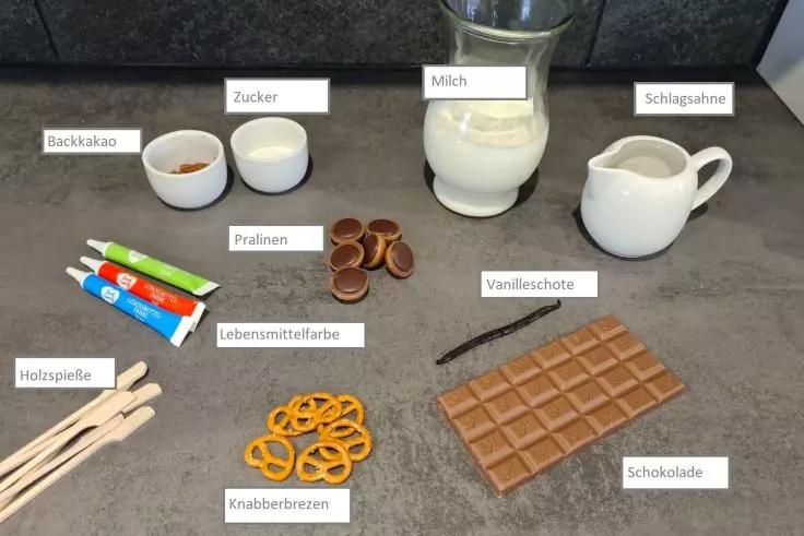 Arbeitsfl&auml;che mit Glas Milch, kleines Milchk&auml;nnchen mit Schlagsahne, zwei kleine Sch&auml;lchen mit Kakaopulver und Zucker, Schokoladentafel, Pralinen, Knabberbrezeln, Tuben Lebensmittelfarbe, Holzspie&szlig;e und Vanilleschote