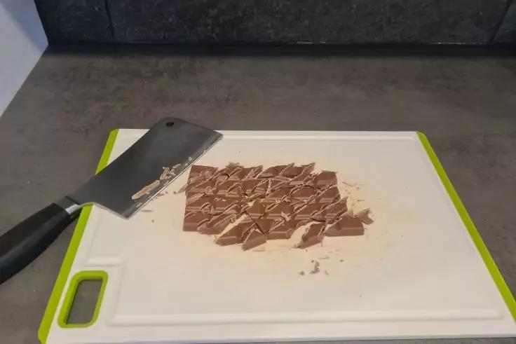 Kleine Schokoladenst&uuml;cke auf einem wei&szlig;en Schneidebrett mit gr&uuml;nem Rand, ein gro&szlig;es Messer mit schwarzem Griff liegt daneben, graue Arbeitsfl&auml;che und dunkle Wand im Hintergrund
