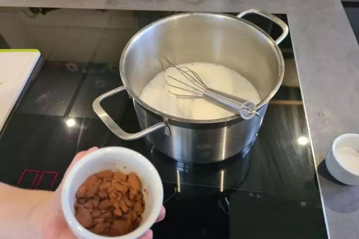 Edelstahl-Topf mit wei&szlig;er Fl&uuml;ssigkeit und Schneebesen auf schwarzem Glaskeramik-Kochfeld; eine Hand h&auml;lt eine kleine wei&szlig;e Schale mit braunen St&uuml;cken daneben; kleine wei&szlig;e Schale rechts auf der Arbeitsfl&auml;che