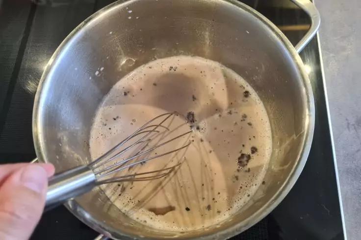 Hand h&auml;lt Schneebesen in einem Metalltopf mit schaumiger hellbrauner Fl&uuml;ssigkeit und dunklen Partikeln auf einer Herdplatte