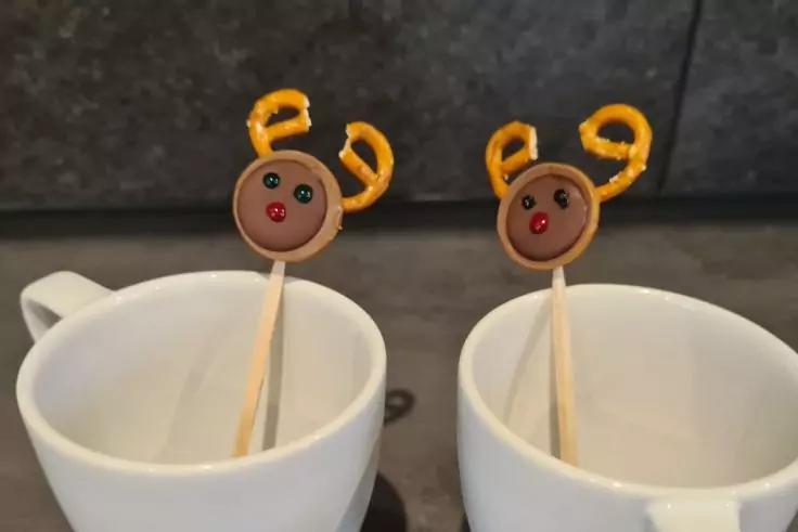 Zwei wei&szlig;e Tassen, jeweils ein Holzstab mit rundem braunem Aufsatz mit zwei schwarzen Punkten, einem roten Punkt und gelben gedrehten Schlaufen