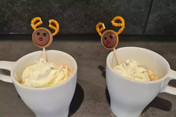 Zwei wei&szlig;e Tassen mit Schlagsahne, jeweils ein Holzst&auml;bchen mit rundem braunem Dekokopf, gelben Geweihen und roter Nase, auf dunkler Arbeitsfl&auml;che