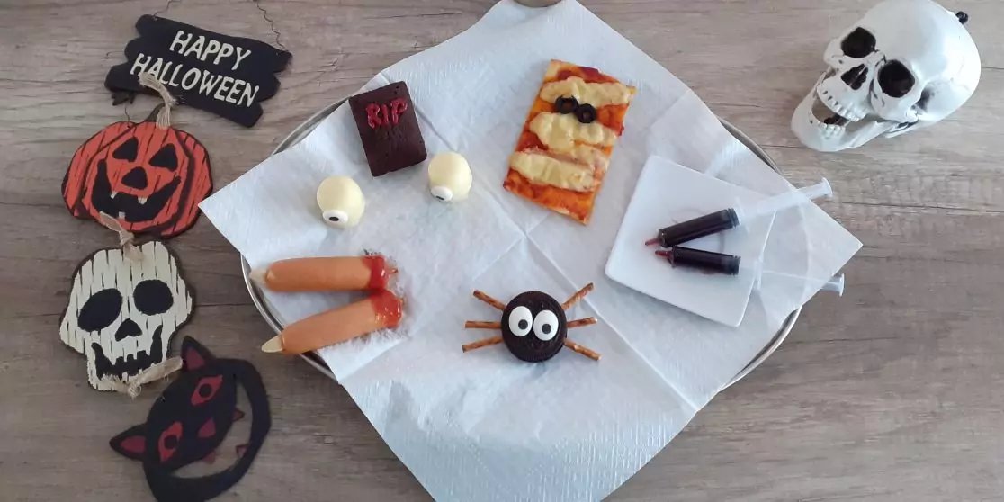 Holztisch mit mehreren Dekorst&uuml;cken links (schwarzes Schild mit Text 'Happy Halloween', orange K&uuml;rbisgesicht, wei&szlig;e Totenkopf- und schwarze Katzen-Auflagen) und einem wei&szlig;en Sch&auml;del rechts. Auf einer wei&szlig;en Serviette in der Mitte: ein rechteckiges Ge