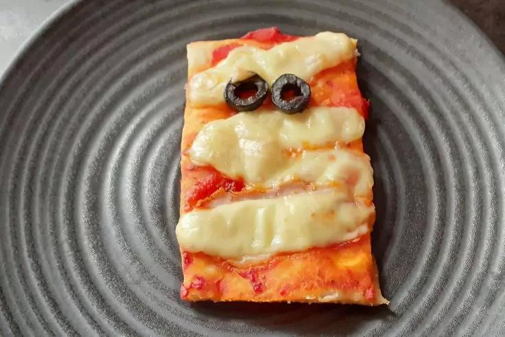 Rechteckiges Pizzast&uuml;ck auf dunklem Teller mit Tomatensauce, drei Streifen geschmolzenem K&auml;se und zwei schwarzen Olivenringen