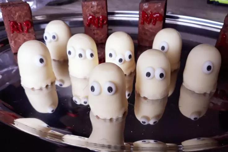 Mehrere wei&szlig;e, zylindrische, mit Schokolade &uuml;berzogene S&uuml;&szlig;igkeiten mit aufgeklebten runden Zuckeraugen auf einem gl&auml;nzenden Metalltablett; Spiegelungen sichtbar; im Hintergrund rote rechteckige Pralinen