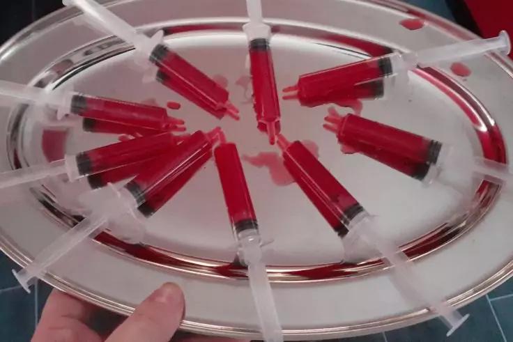 Rundes Metalltablett mit radial angeordneten Spritzen, jede mit roter Fl&uuml;ssigkeit, gehalten von einer Hand