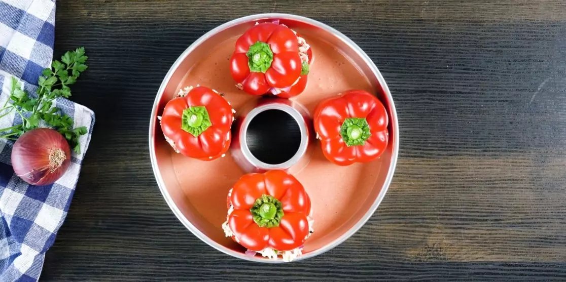 Draufsicht auf vier rote Paprika in einer runden Form um eine kleine Schale mit dunkler Fl&uuml;ssigkeit, auf dunklem Holztisch; links kariertes K&uuml;chentuch, rote Zwiebel und Petersilie