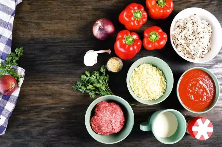 Schalen und Gegenstände auf dunklem Holztisch: drei rote Paprika, weiße Schale mit Reis, rote Zwiebel, Knoblauchzehe, kleine Schale mit Gewürz, Petersilie, grüne Schalen mit geriebenem Käse, rohes Hackfleisch, Tomatensauce, kleine Milchkanne, rot-wei