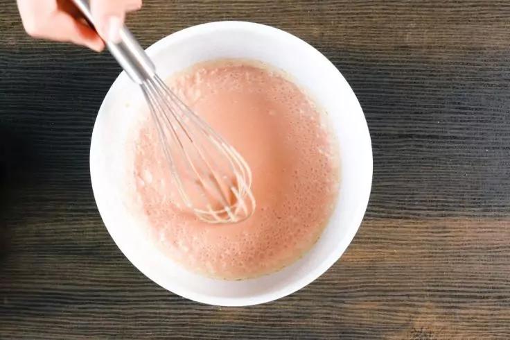 Weiße Schüssel mit rosa Flüssigkeit, Metallbesen in der Flüssigkeit, Hand hält den Besen oben links, dunkle Holzoberfläche darunter